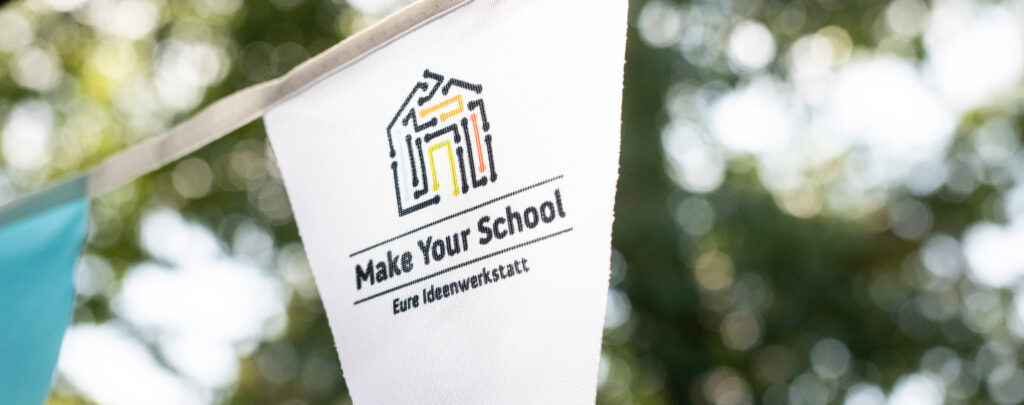 Das Foto zeigt eine Nahaufnahme von einem weißen Wimpel, der an einer Girlande befestigt ist. Darauf ist das Make Your School Logo abgebildet. Im Hintergrund sind verschwommen Bäume zu erahnen.