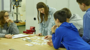 Eine junge Frau beugt sich über Schüler und schaut ihnen über die Schulte wie sie Karten mit technischen Bauteilen darauf begutachten.