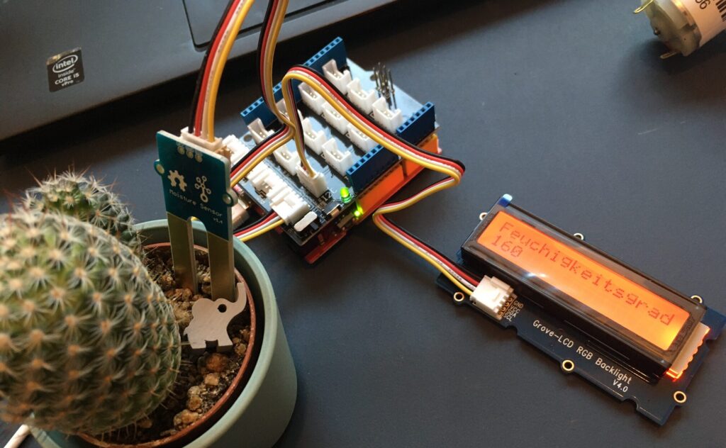 Links ist eine Kaktuspflanze, in den Boden gesteckt ist ein Feuchtigkeitssensor. Der Sensor ist verbunden mit einem Arduino UNO und einem Bildschirm, auf dem steht: "Feuchtigkeitsgrad 160". 