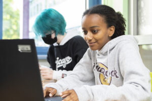 Im Vordergrund sitzt ein Mädchen an einem Laptop. Im Hintergrund bastelt ein Mädchen konzentriert.