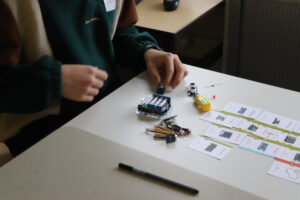 Auf einem Tisch liegen Materialkarten mit Abbildungen von elektronischen Bauteilen. Ein Jugendlicher hält einen Mikrocontroller in der Hand.