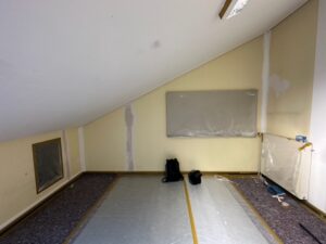 Ein leerer Raum mit schräger Decke. Die Wände sind hellgelb. Der Boden ist mit Folie und Malerflies ausgelegt.