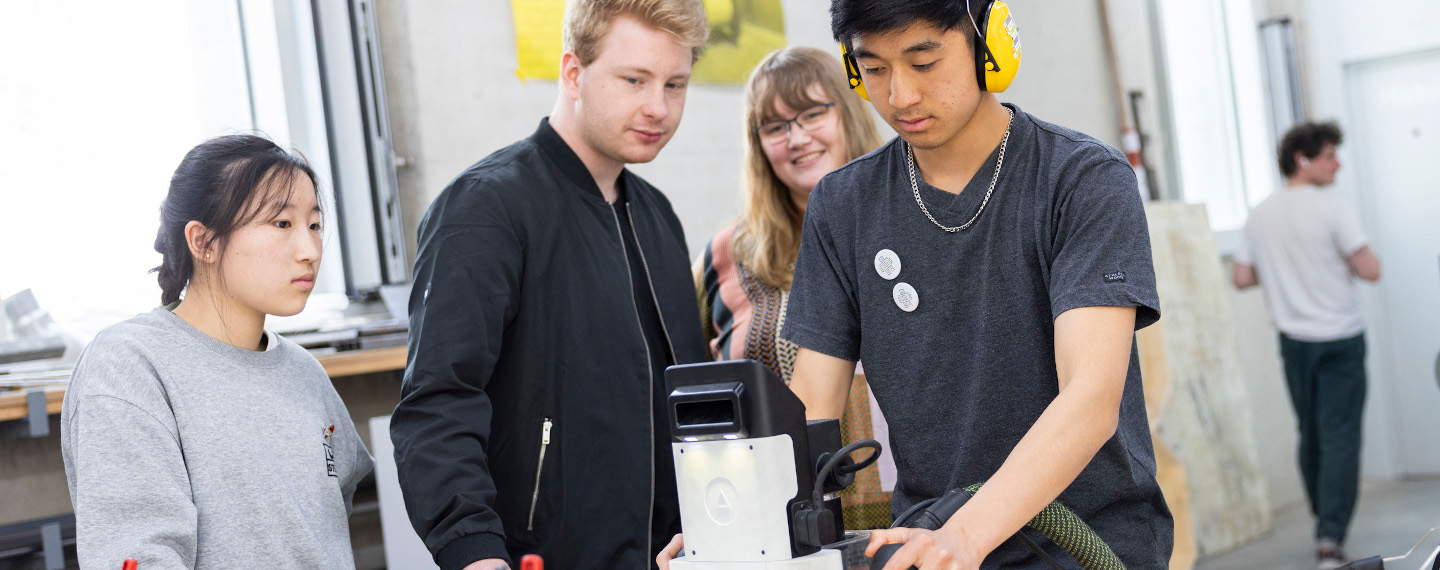 Ein Jugendlicher steht mit Kopfhörern in einem Makerspace und bedient eine Fräse. Um ihn herum stehen eine weitere Jugendliche und zwei Mentor*innen.