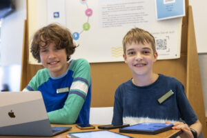 Zwei Jungs sitzen an einem Tisch und grinsen in die Kamera. Vor ihnen liegen zwei Handys, zwei Tablets und ein Laptop. Hinter ihnen ist eine Stellwand mit einem Poster daran zu erkennen.