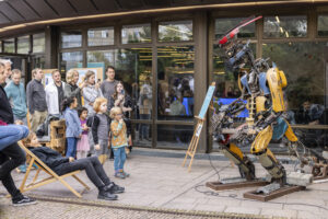 Auf der rechten Seite steht ein etwa dreieinhalb Meter großer, metallischer Roboter. Er ist aus grauen, blauen und gelben Schrotteilen zusammengebaut. Mit langen, dünnen Fingern spielt er auf einer Bassgitarre. Auf der linken Seite stehen kleine Kinder mit ihren Eltern und schauen dem Roboter fasziniert zu.