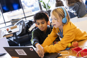 Zwei Kinder arbeiten gemeinsam an einem Laptop. Beide tragen Kopfhörer. Vor ihnen liegen einige Äpfel, in die sie Drähte gesteckt haben. Sie tippen zusammen mit drei Händen gleichzeitig auf der Tastatur.